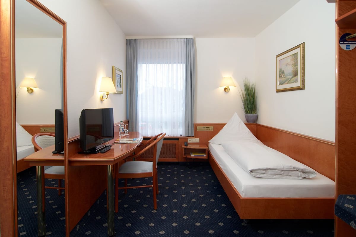 A hotel room with a bed, a desk with a chair, a television, a lamp, a plant, and a window with curtains.