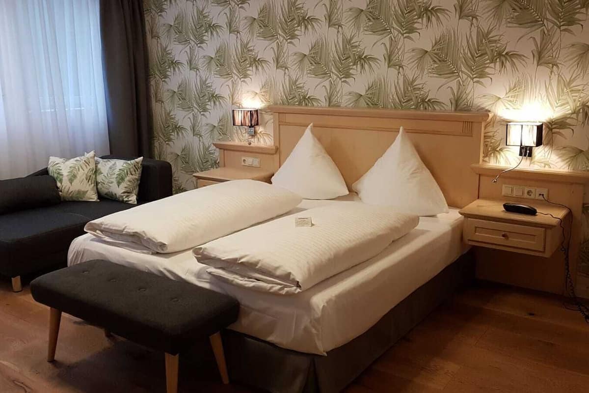 A double bed with two pillows, a bedside table with a lamp, a small bedside table with a lamp, a dark-colored sofa, and a bench.