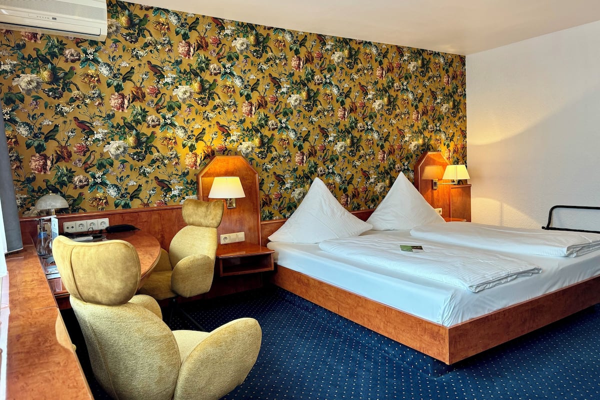 A hotel room with a floral wallpaper, a bed with white linens, two bedside lamps, a wooden nightstand, a chair, and a desk with a lamp.