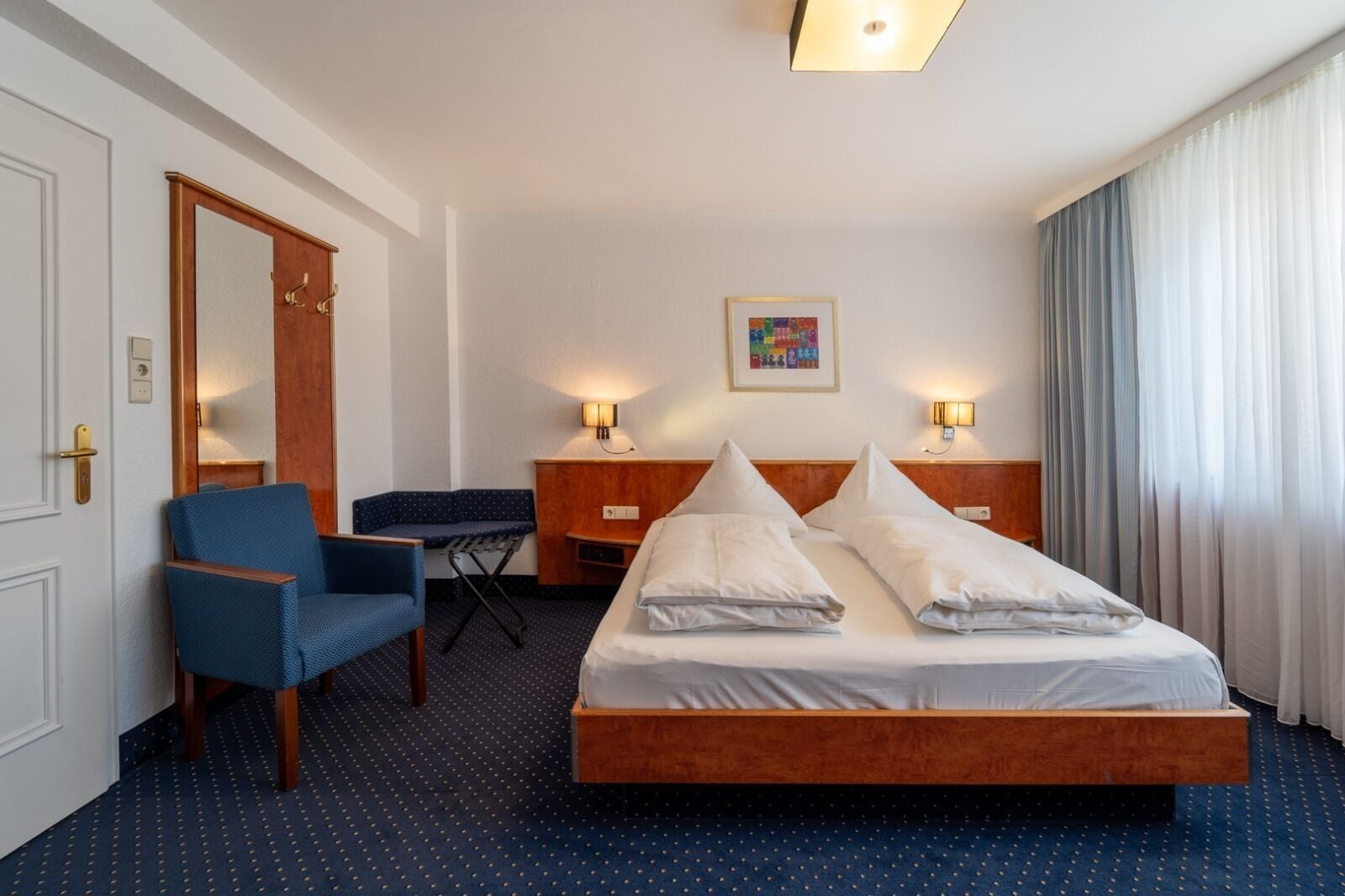 Habitación de hotel con cama, dos mesitas de noche con lámparas, una silla, un escritorio, un espejo y un cuadro en la pared.