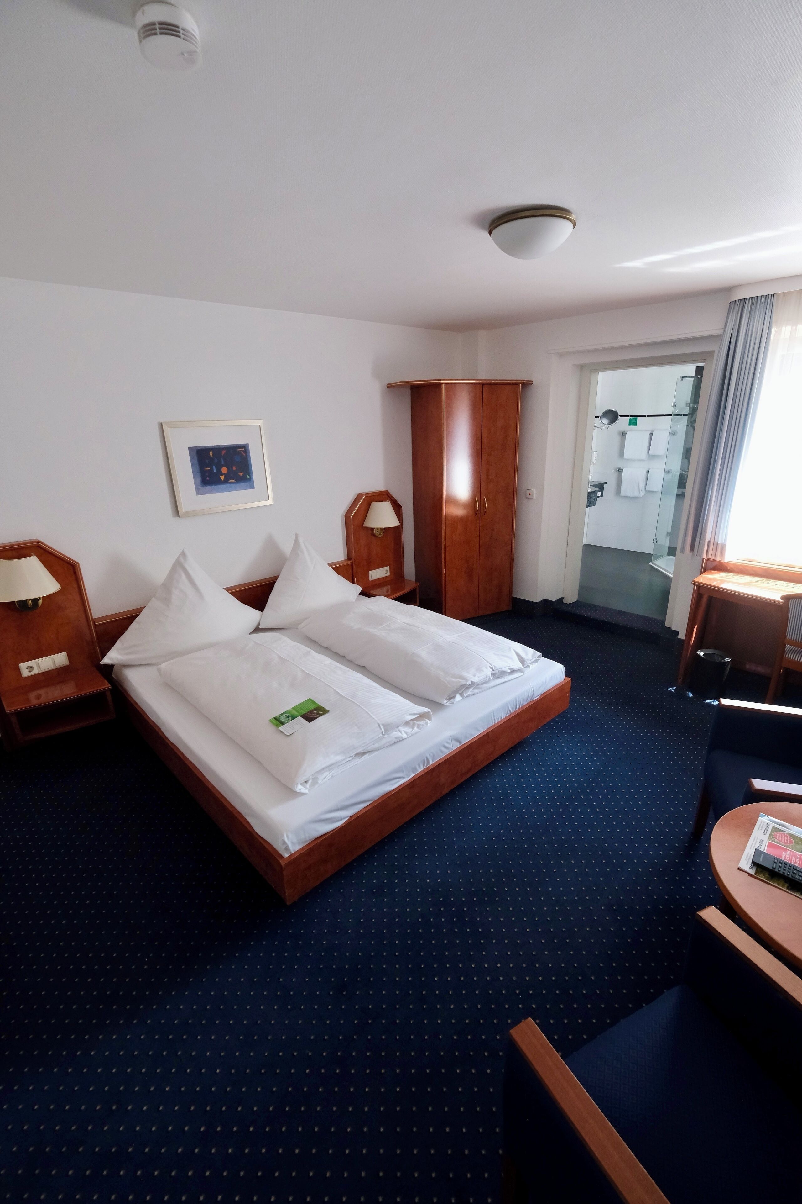Une chambre d’hôtel comprenant un lit, un bureau, une armoire et une salle de bain visible à travers une porte en verre.
