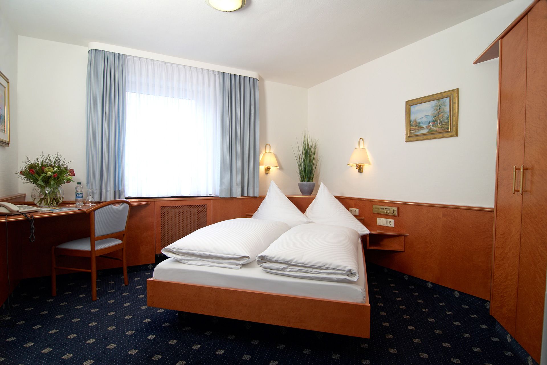 A hotel room with a bed, a desk, a chair, a window with curtains, a plant, and a painting on the wall.