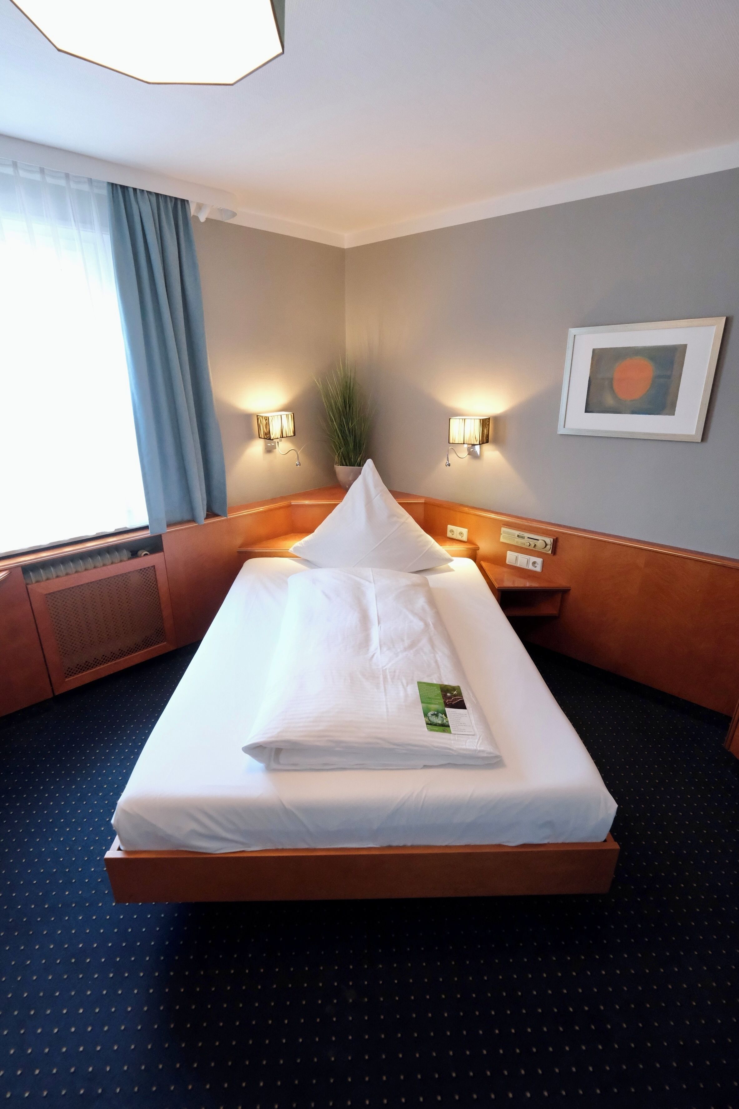 A hotel room with a bed, two bedside lamps, a framed picture on the wall, and a window with curtains.