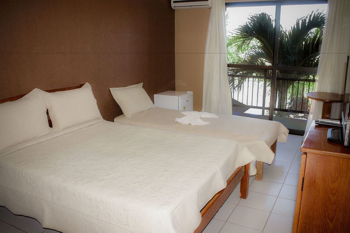 A hotel room with two beds, a wooden headboard, a small bedside table, a desk with a computer, and a view of palm trees through a balcony.