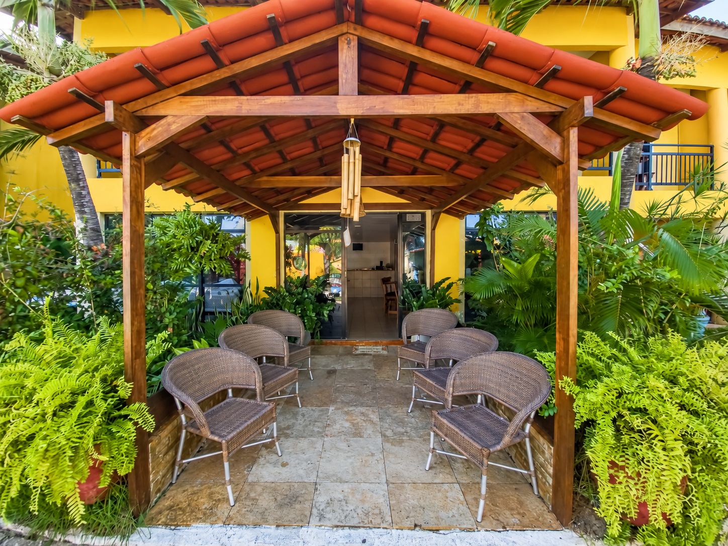 Un espacio al aire libre con techo, con sillas de mimbre y suelo de baldosas, rodeado de abundante vegetación.