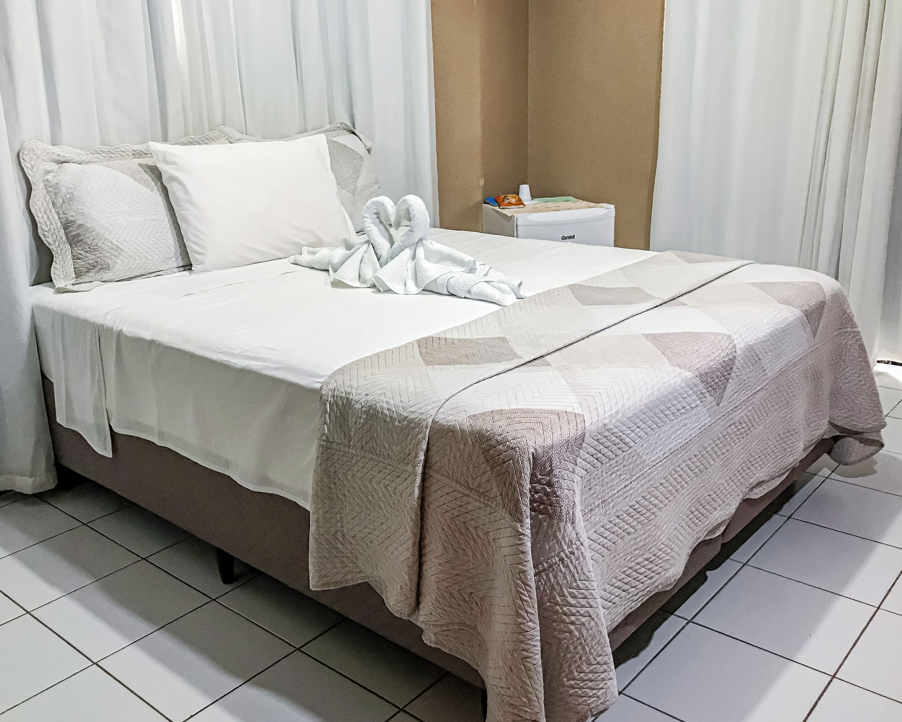 Cama bem arrumada com edredom branco, colcha bege e toalha dobrada em formato de coração.