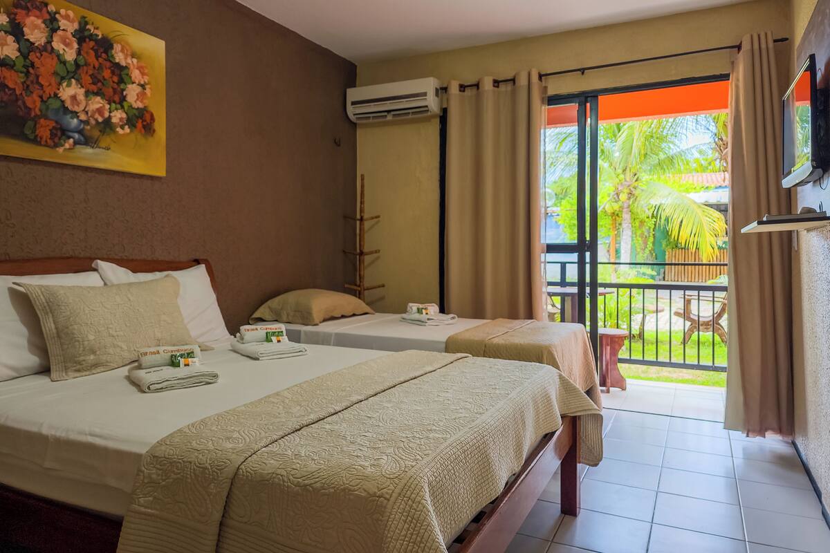 A hotel room with two beds, a painting on the wall, an air conditioning unit, and a balcony with a view of greenery.
