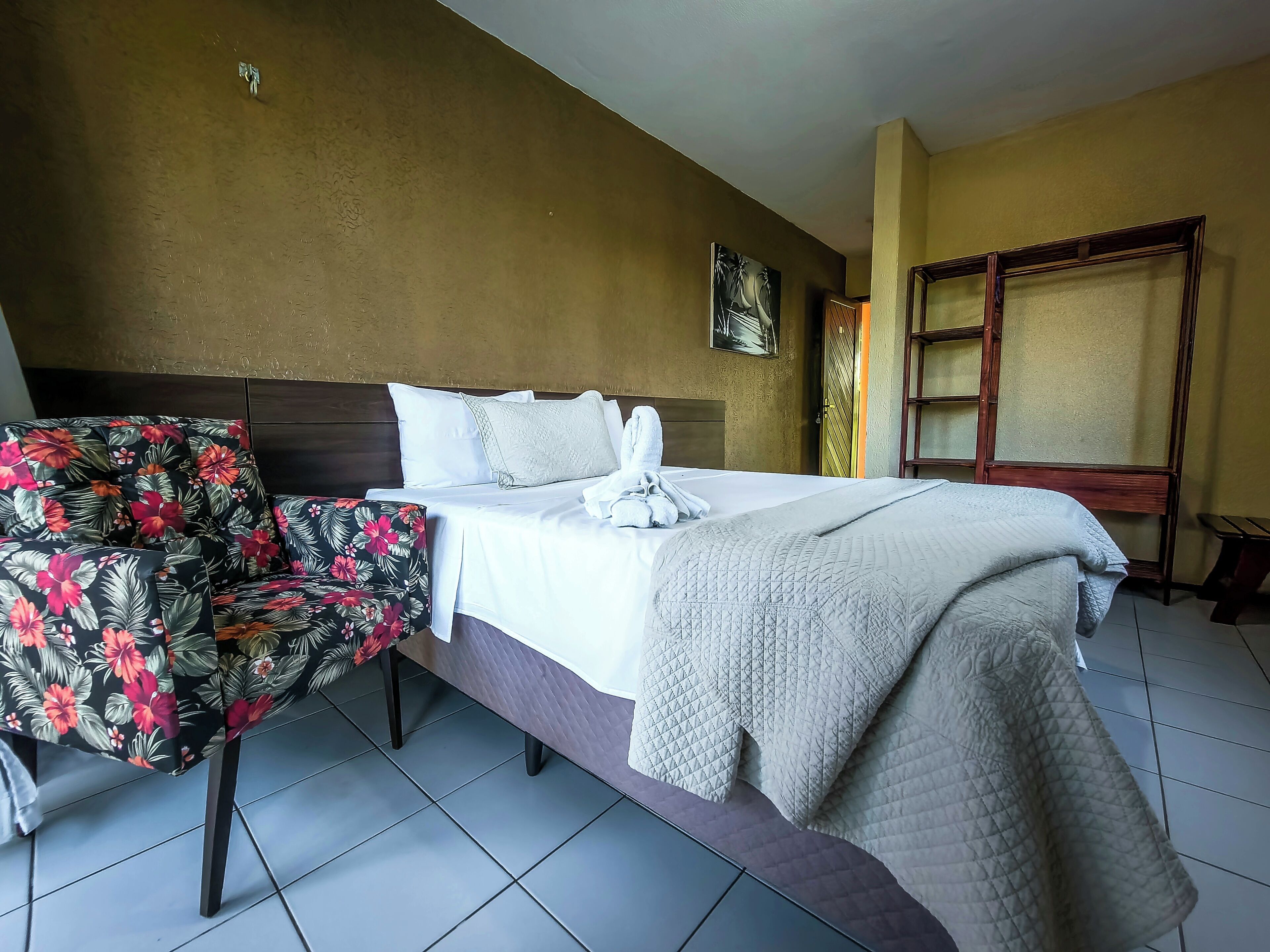 A hotel room with a bed, a floral chair, a painting on the wall, and a window with a view.