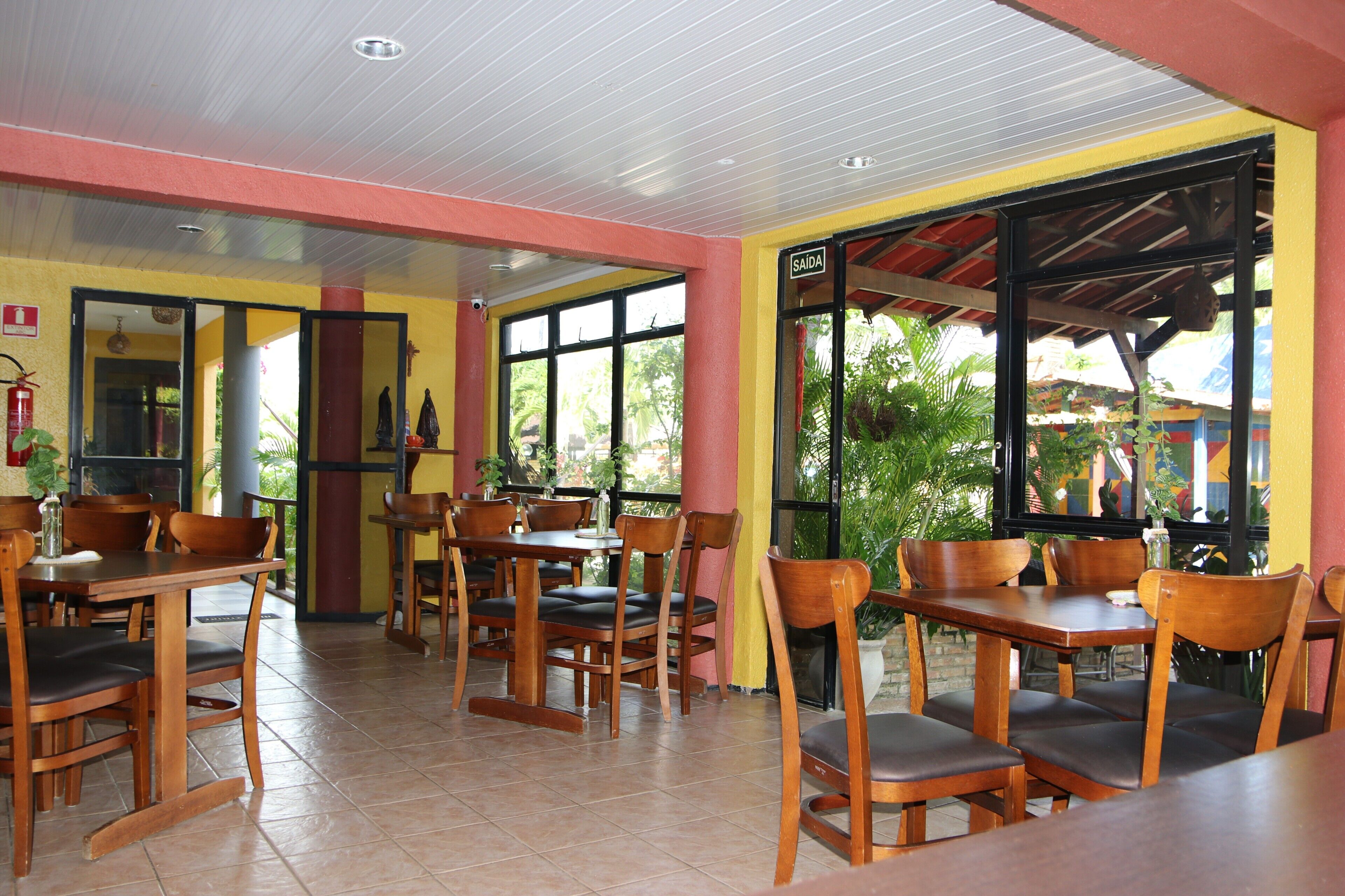 An indoor dining area with wooden tables and chairs, large windows with a view of greenery, and a red pillar.