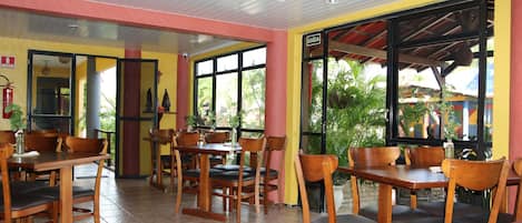 An indoor dining area with wooden tables and chairs, large windows with a view of greenery, and a red pillar.