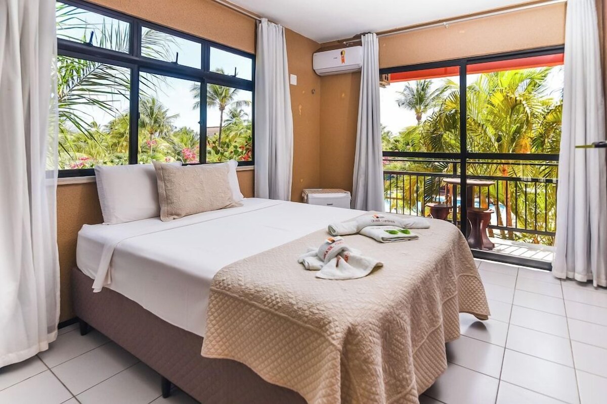A hotel room with a bed, a balcony with a view of palm trees, and an air conditioning unit.