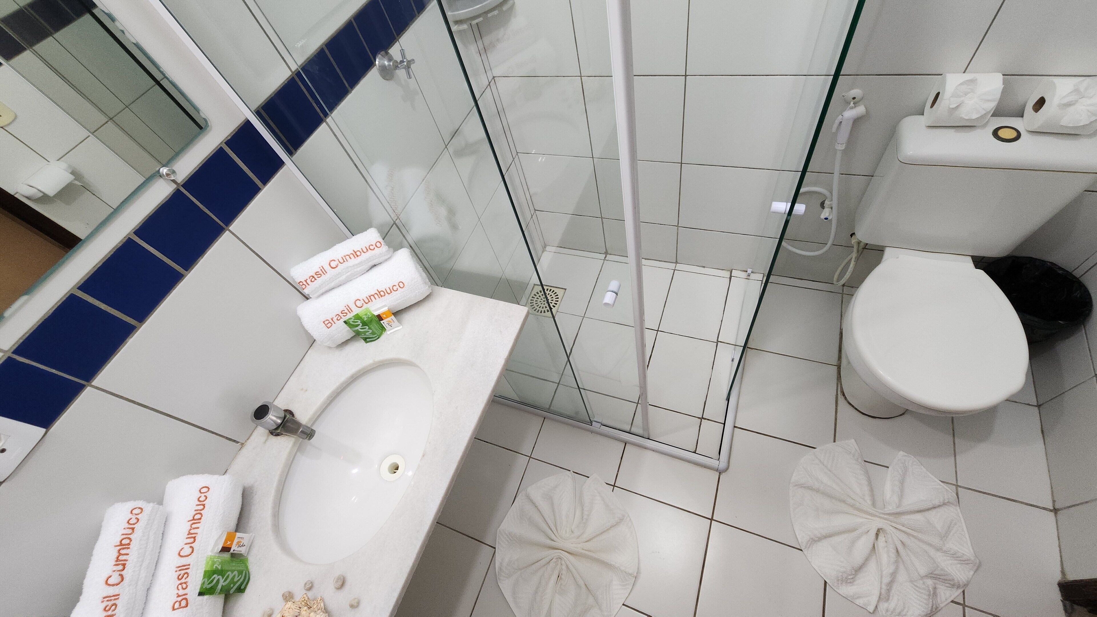 A bathroom with a white sink, a toilet, and a glass shower enclosure. There are two white towels with "Brasil Cumbuco" on them.