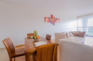 A dining area with a wooden table, chairs, and a fruit bowl. There is a white sofa and a balcony with a view. - Lymack Suites (Nairobi)