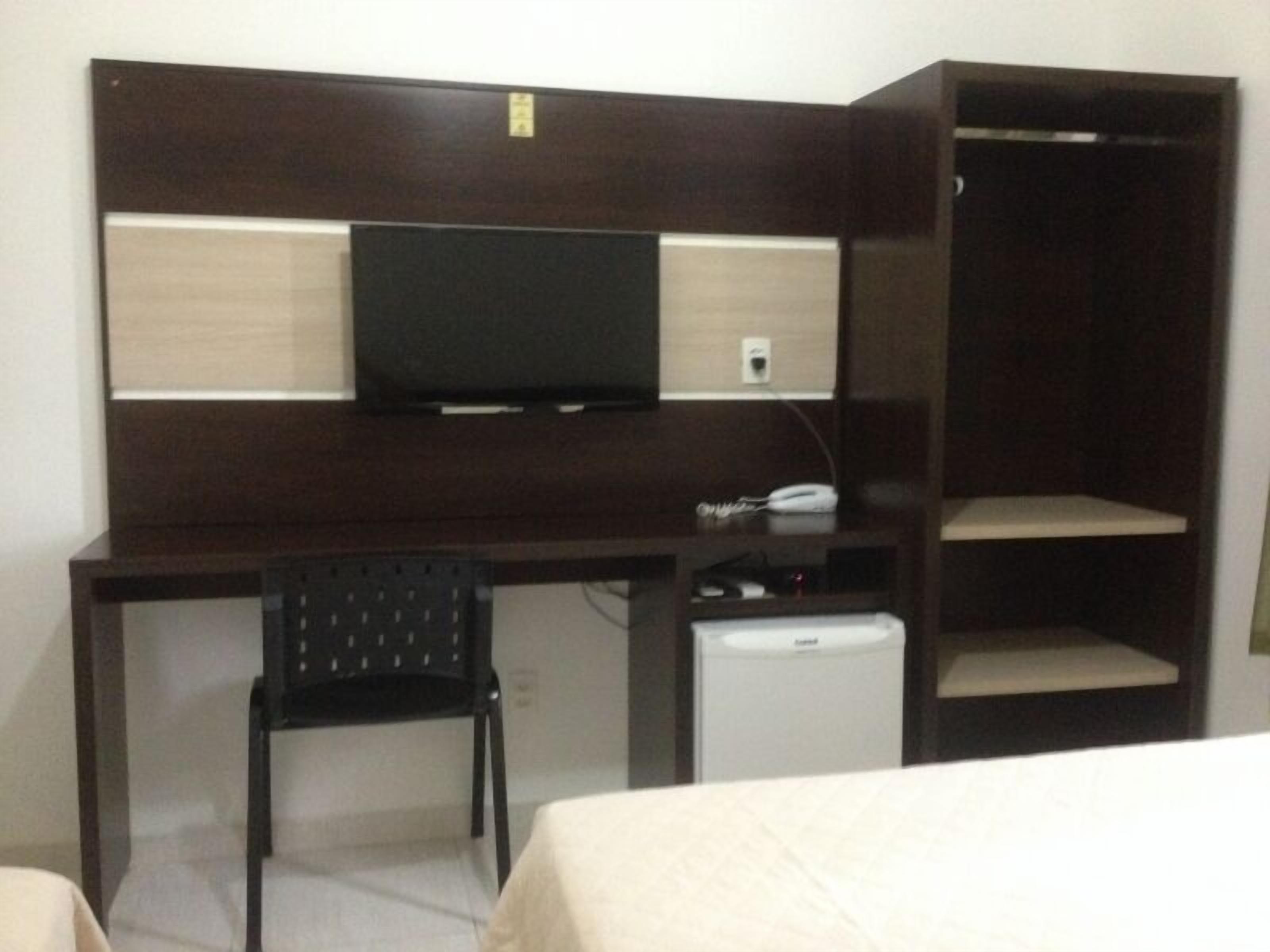 Habitación de hotel compacta con un escritorio de madera oscura, una pequeña silla negra, un televisor y un mini refrigerador.
