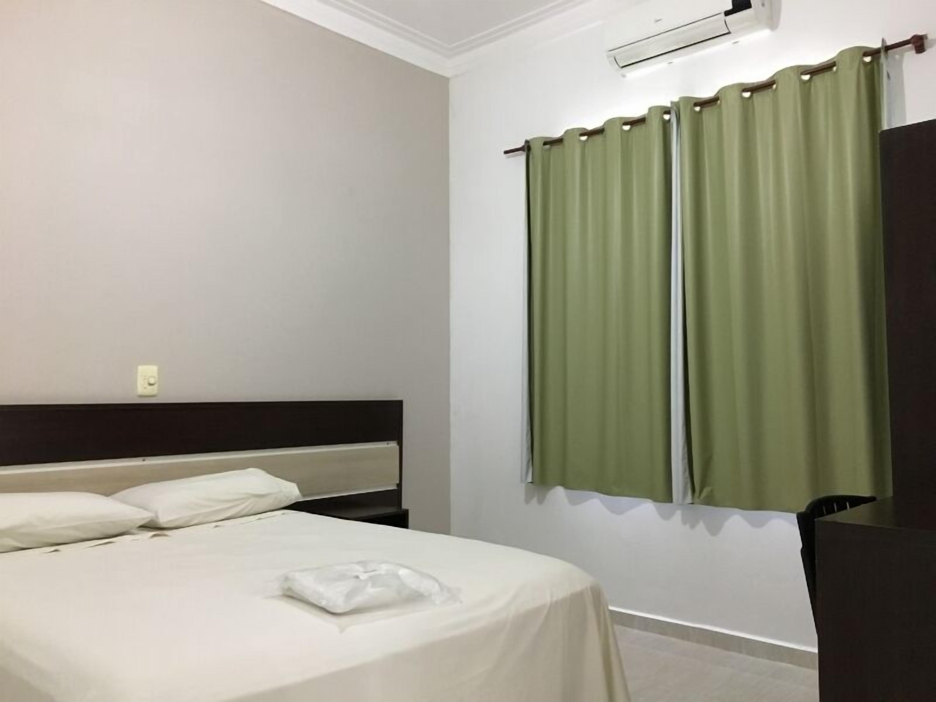 Habitación de hotel con cama, cabecera, aire acondicionado de pared y cortinas verdes.