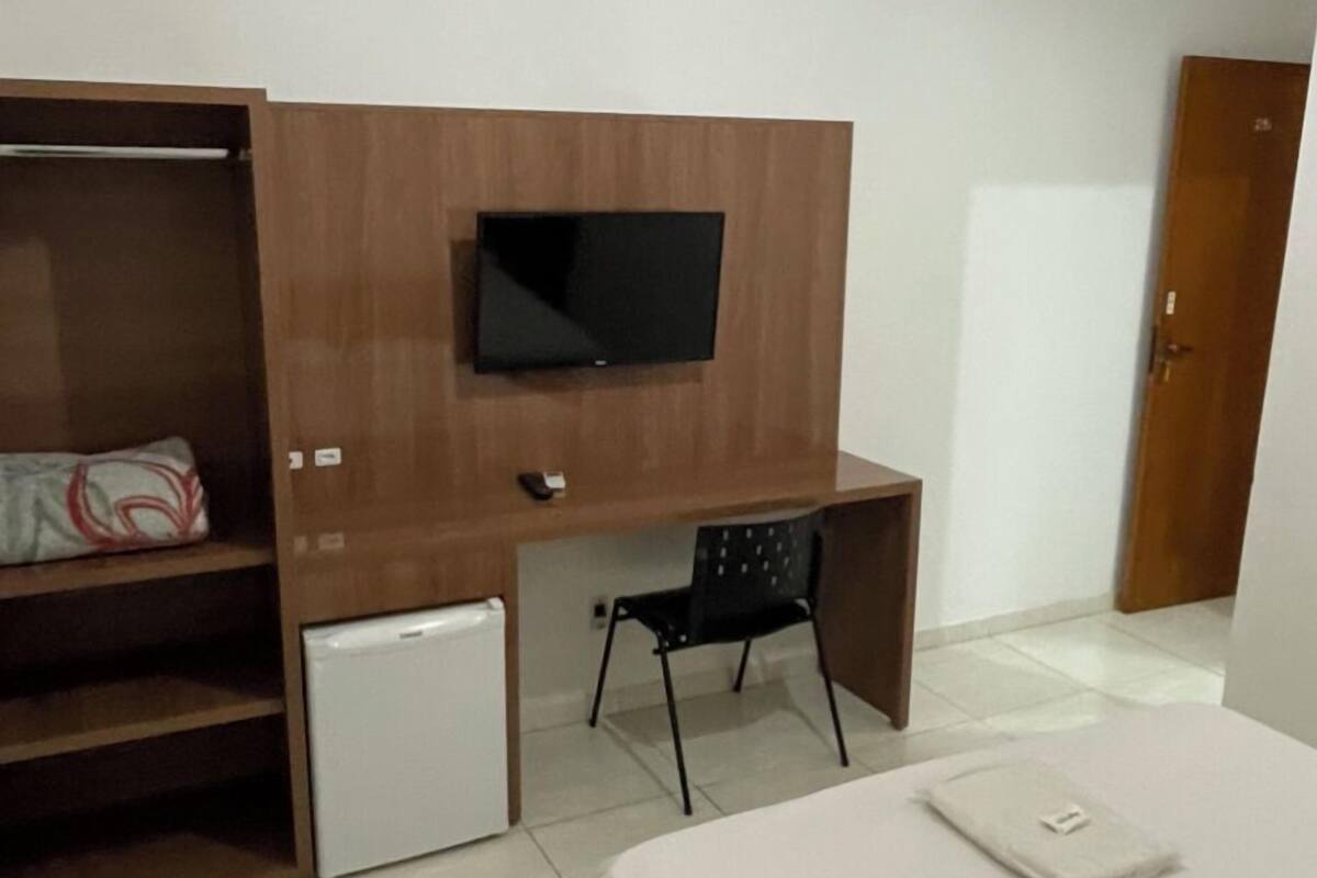 A bedroom with a bed, a desk, a TV mounted on a wooden cabinet, a small fridge, and a wardrobe.
