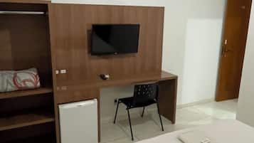 A bedroom with a bed, a desk, a TV mounted on a wooden cabinet, a small fridge, and a wardrobe.