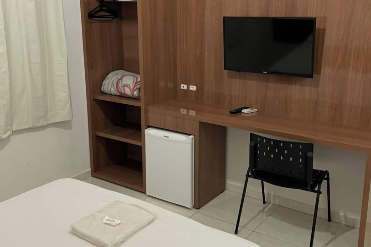 A bedroom with a bed, a desk with a TV, a mini fridge, a chair, and a wardrobe.