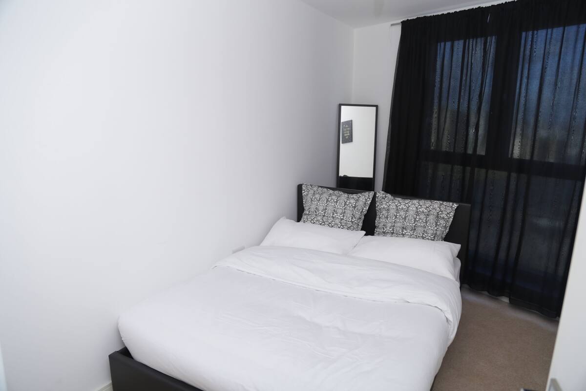 A neatly made bed with white linens and patterned pillows, a bedside mirror, and a window with dark curtains.