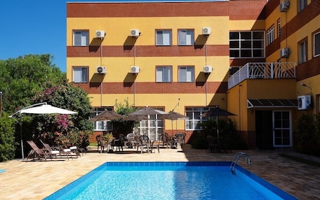 A swimming pool with lounge chairs and umbrellas, a multi-story building with air conditioning units, and a clear blue sky.