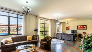 A spacious living room with a large sofa, armchair, and coffee table. There are two large windows with curtains, a television, and a painting on the wall. - Itapetinga Hotel (Atibaia)