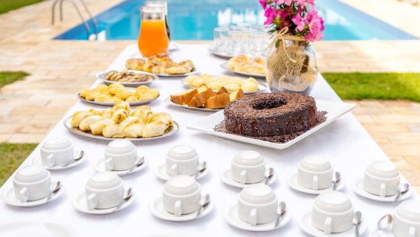 A breakfast buffet with various pastries, a chocolate cake, and white cups on a table by a pool. - Itapetinga Hotel (Atibaia)