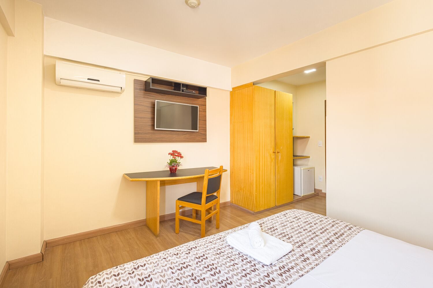 A hotel room with a bed, a desk with a chair, a TV mounted on the wall, and a wardrobe.
