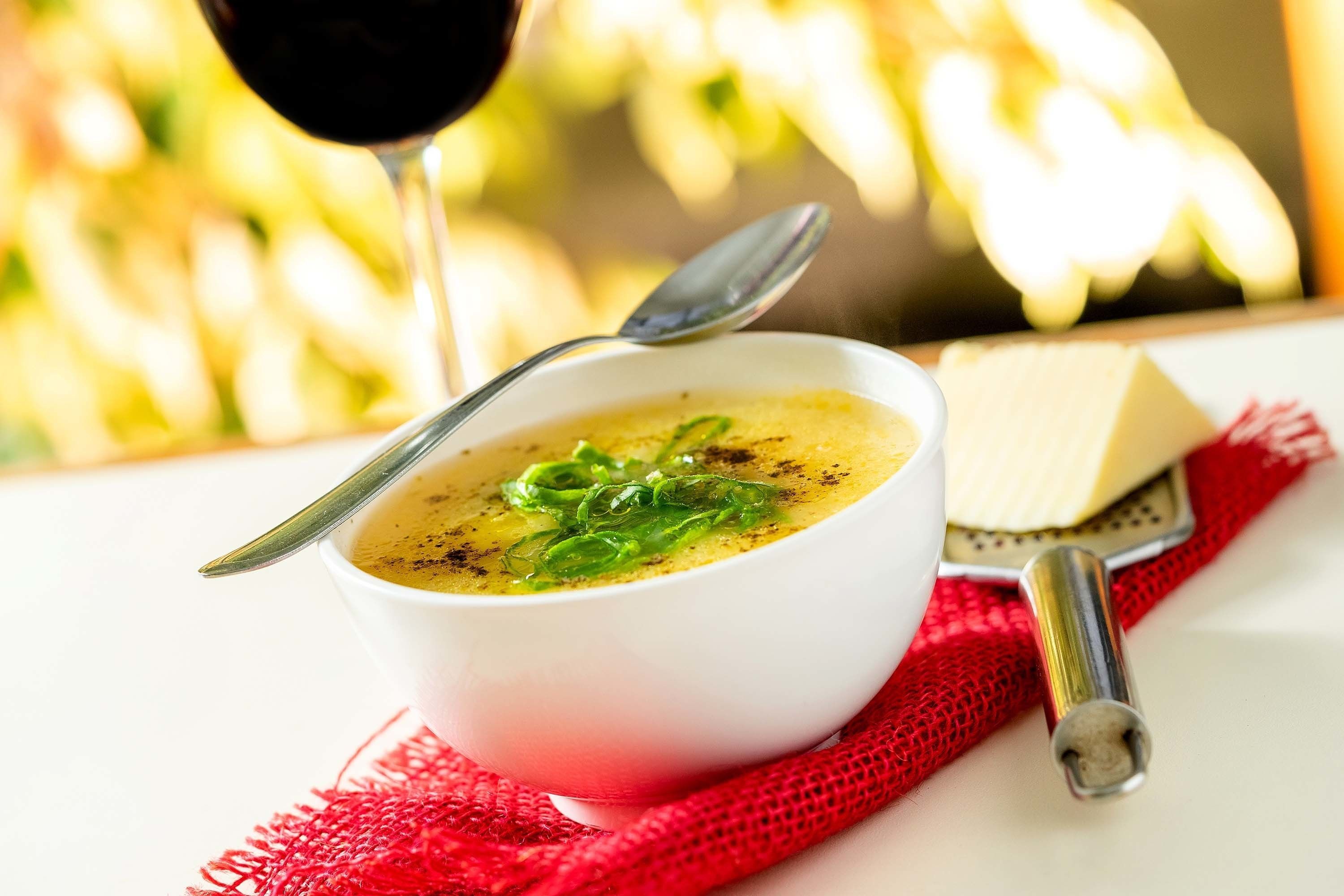 Un tazón de sopa con cuchara, una copa de vino tinto y un trozo de queso sobre un mantel rojo.
