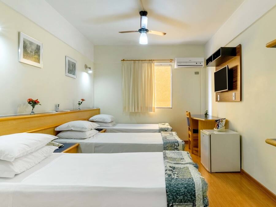 Hotelkamer met drie bedden, een plafondventilator, een airconditioner, een aan de muur bevestigde televisie en een kleine koelkast.