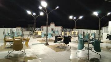 Una terraza en la azotea con varias sillas y mesas, iluminada por luces colgantes.