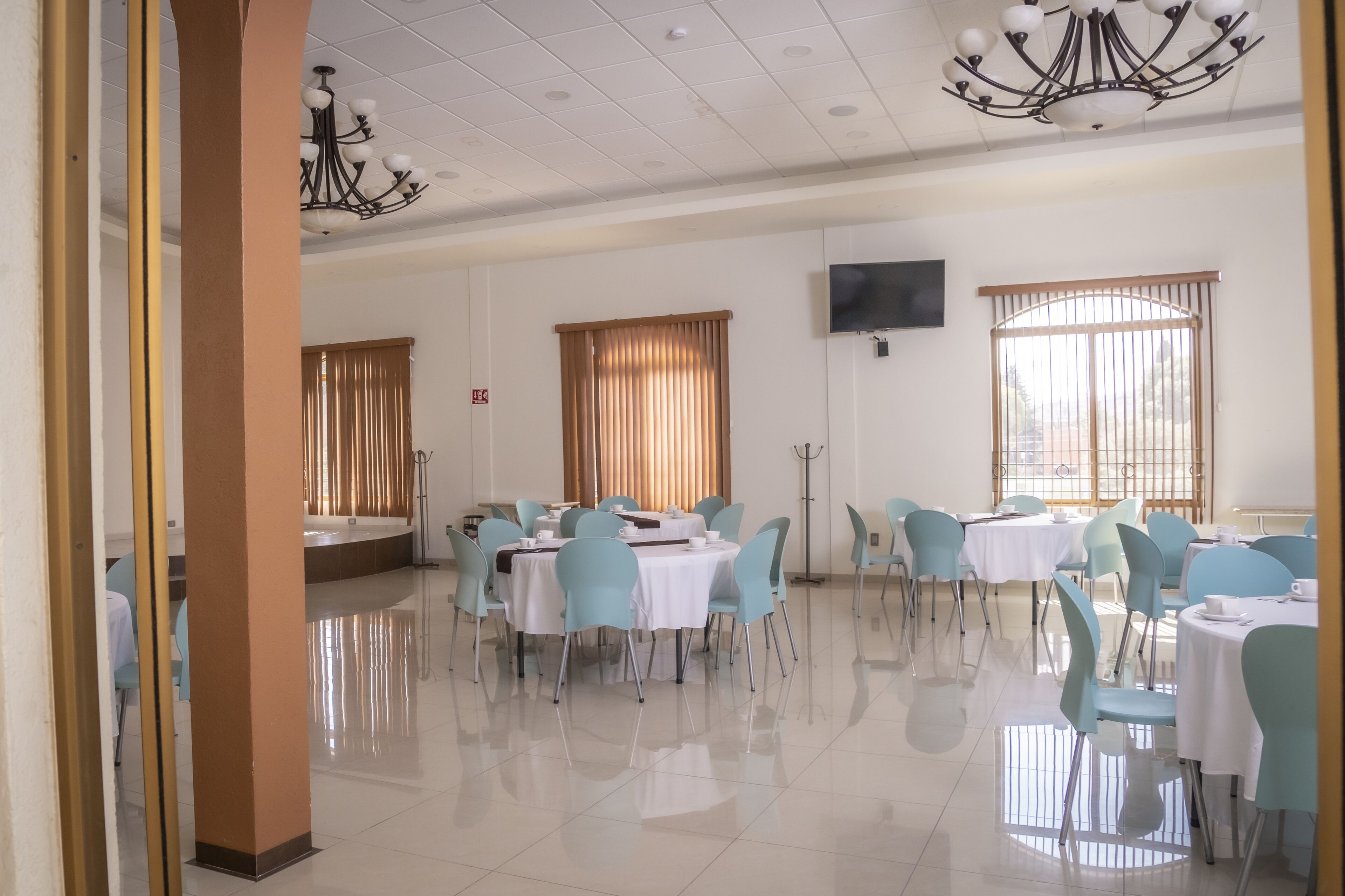 A spacious dining area with round tables and light blue chairs, large windows with blinds, and a chandelier.