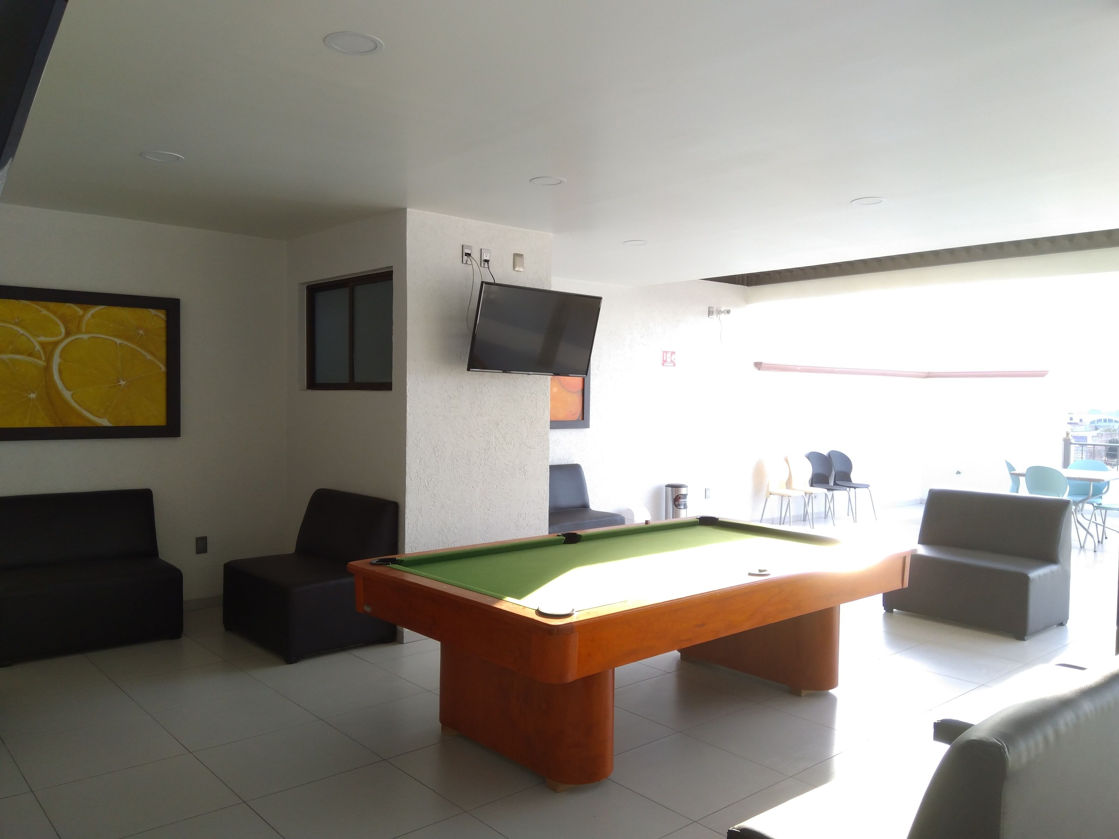 Uma mesa de bilhar em um ambiente bem iluminado, com uma televisão na parede e uma pintura grande de laranjas.