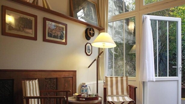 A cozy room with a wooden bench, two striped chairs, a table with a lamp, and a large window with a view of greenery. - Les Chambres de Mathilde (Angers)