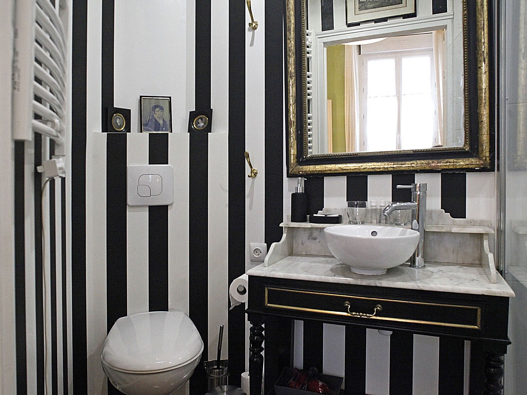 Een badkamer met een wit toilet, een zwart-wit gestreepte wand, een witte wastafel met gouden kraan en een spiegel met gouden lijst.