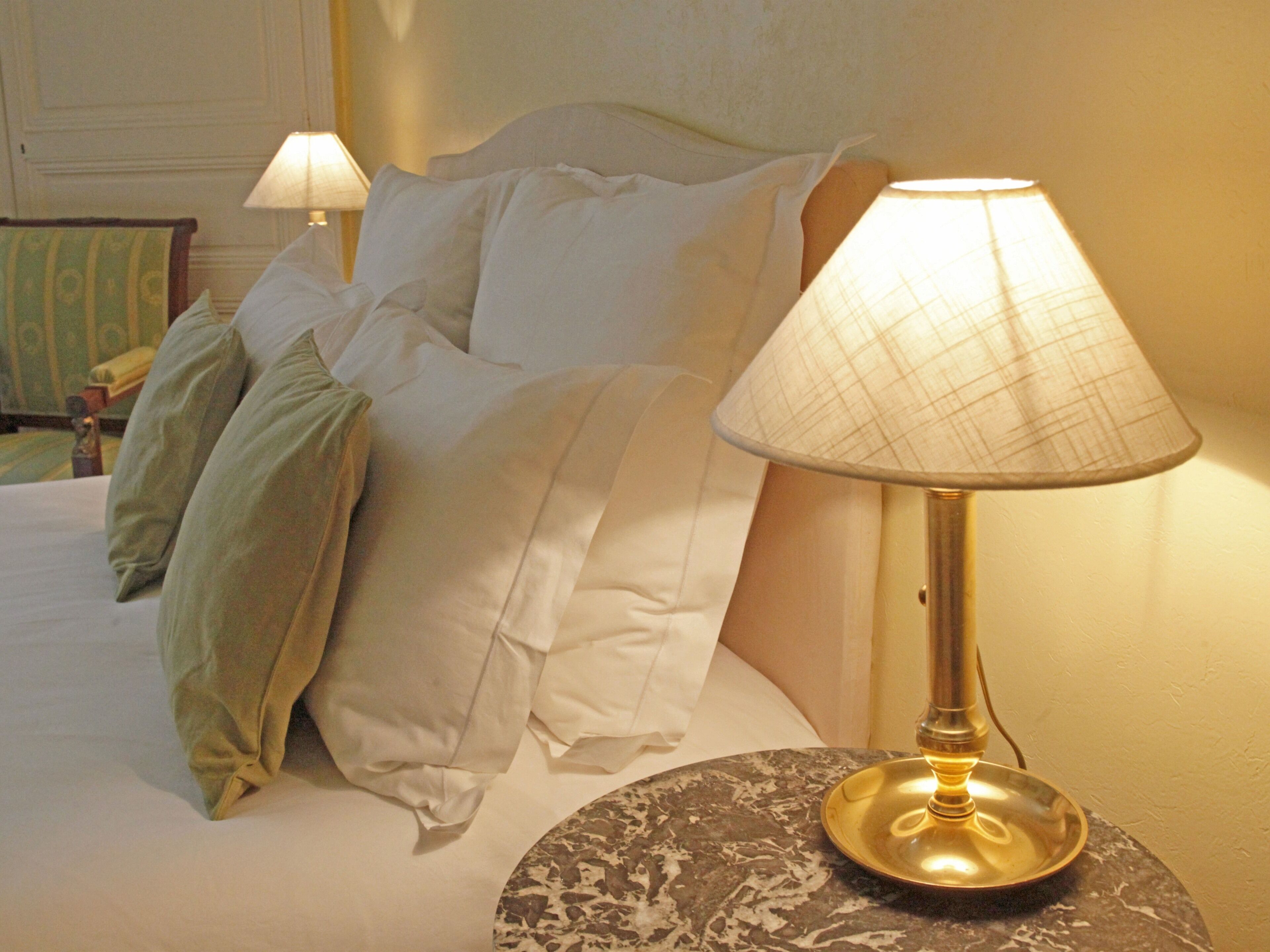 Een net opgemaakt bed met witte lakens en een gouden lamp op een rond nachtkastje.