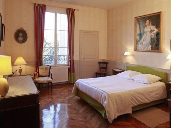 A bedroom with a bed, two chairs, a desk, a window with curtains, a radiator, and a painting on the wall. - Les Chambres de Mathilde (Angers)