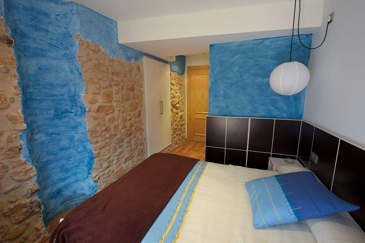 Ein Schlafzimmer mit einer Steinwand, einem Bett mit blau-gelb gestreifter Bettdecke und einer weißen Hängelampe.