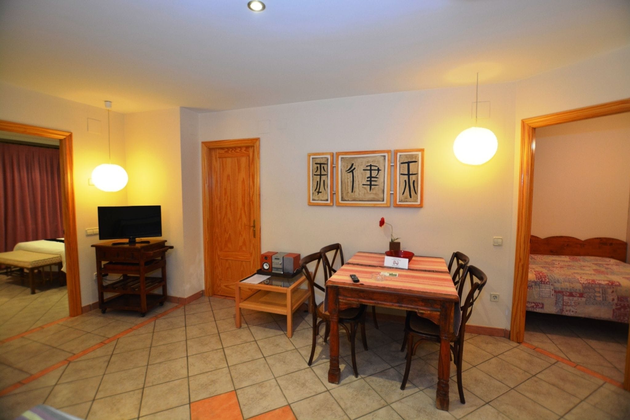 Una habitación con una mesa de comedor de madera, sillas, televisor, un escritorio pequeño y una cama con colcha.