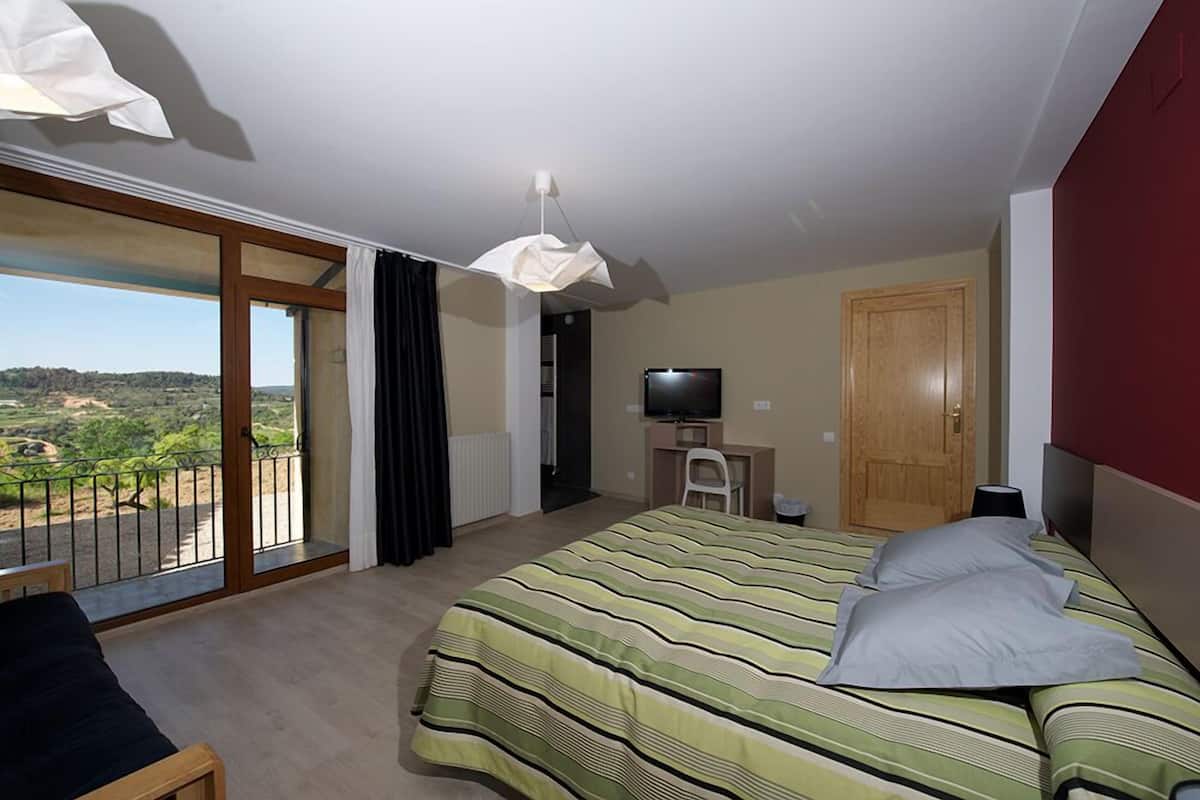 Ein Schlafzimmer mit einem großen Bett, einem Schreibtisch mit Computer, einem Fernseher und einem Balkon mit Blick auf die Landschaft.