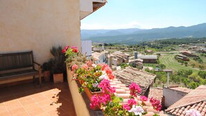 Un balcón con un banco y plantas en macetas, con vistas a un pueblo con campos en terrazas y montañas al fondo.