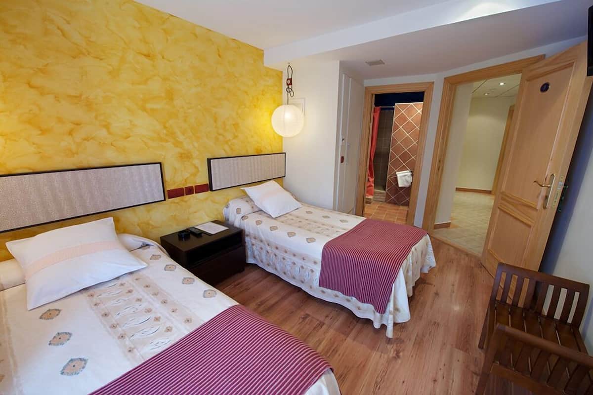 Ein Hotelzimmer mit zwei Betten, Holzboden, gelber Wand mit Struktur, Nachttisch mit Lampe und Holzstuhl.