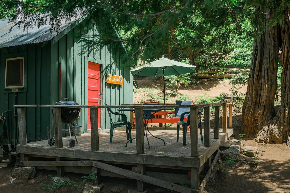 A wooden deck with a grill and chairs, a red door, and a green umbrella.