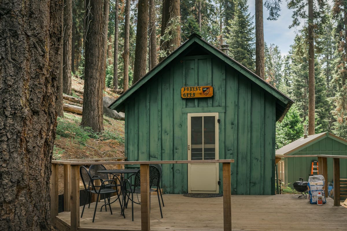 A green cabin with a sign reading "Forest Cabin" and a small deck with chairs.