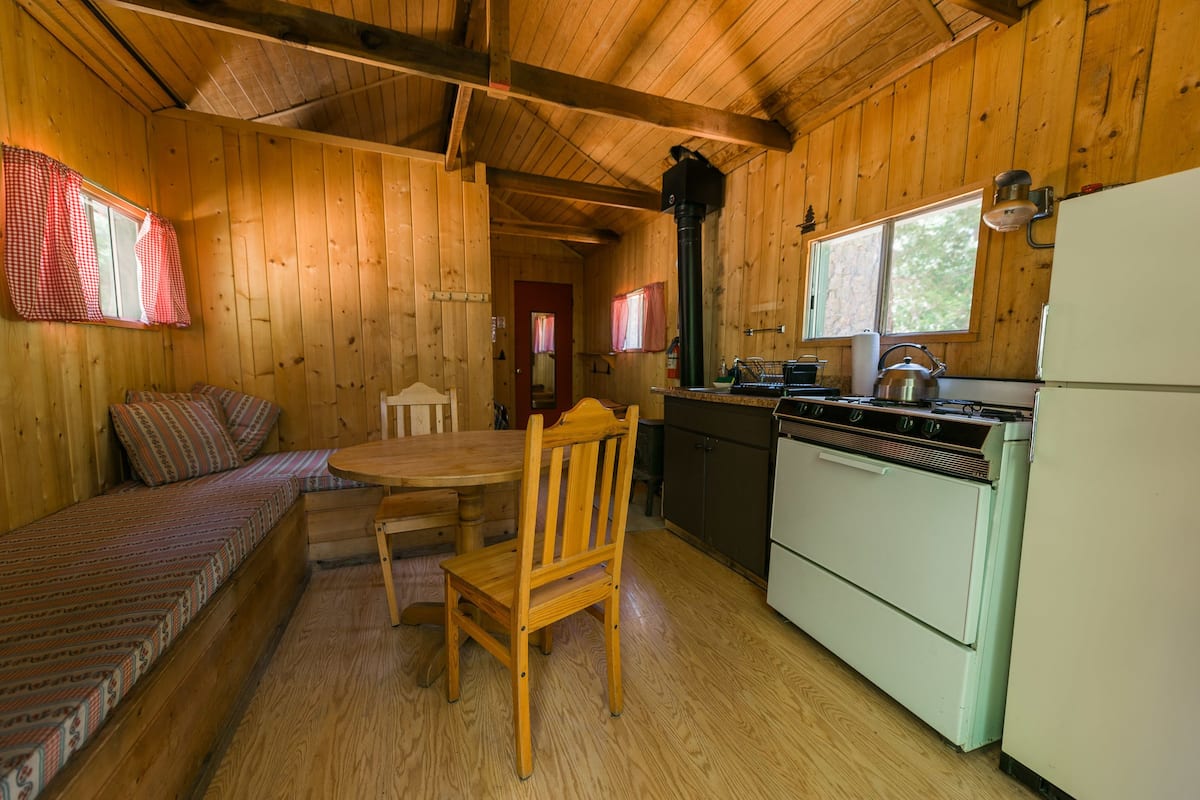 A wooden cabin interior with a dining area, a kitchen with a stove and refrigerator, and a window with curtains.