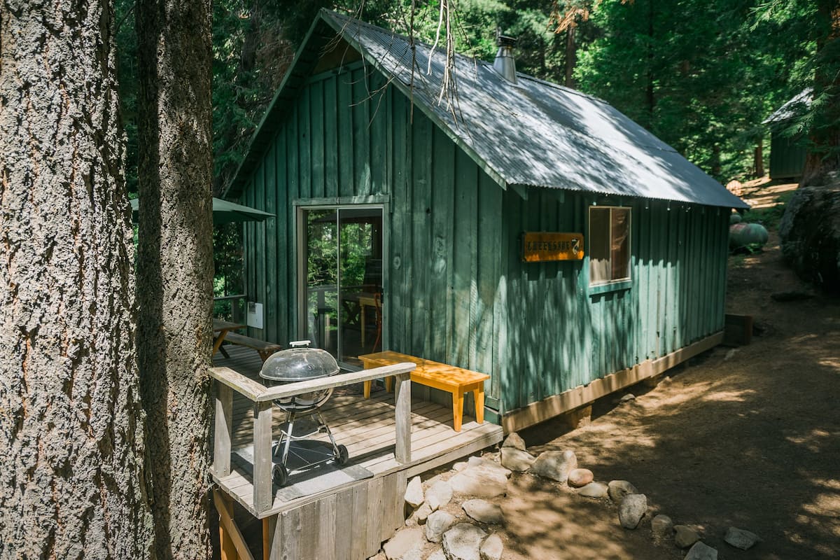 A small, rustic cabin with a wooden deck, a grill, and a bench.