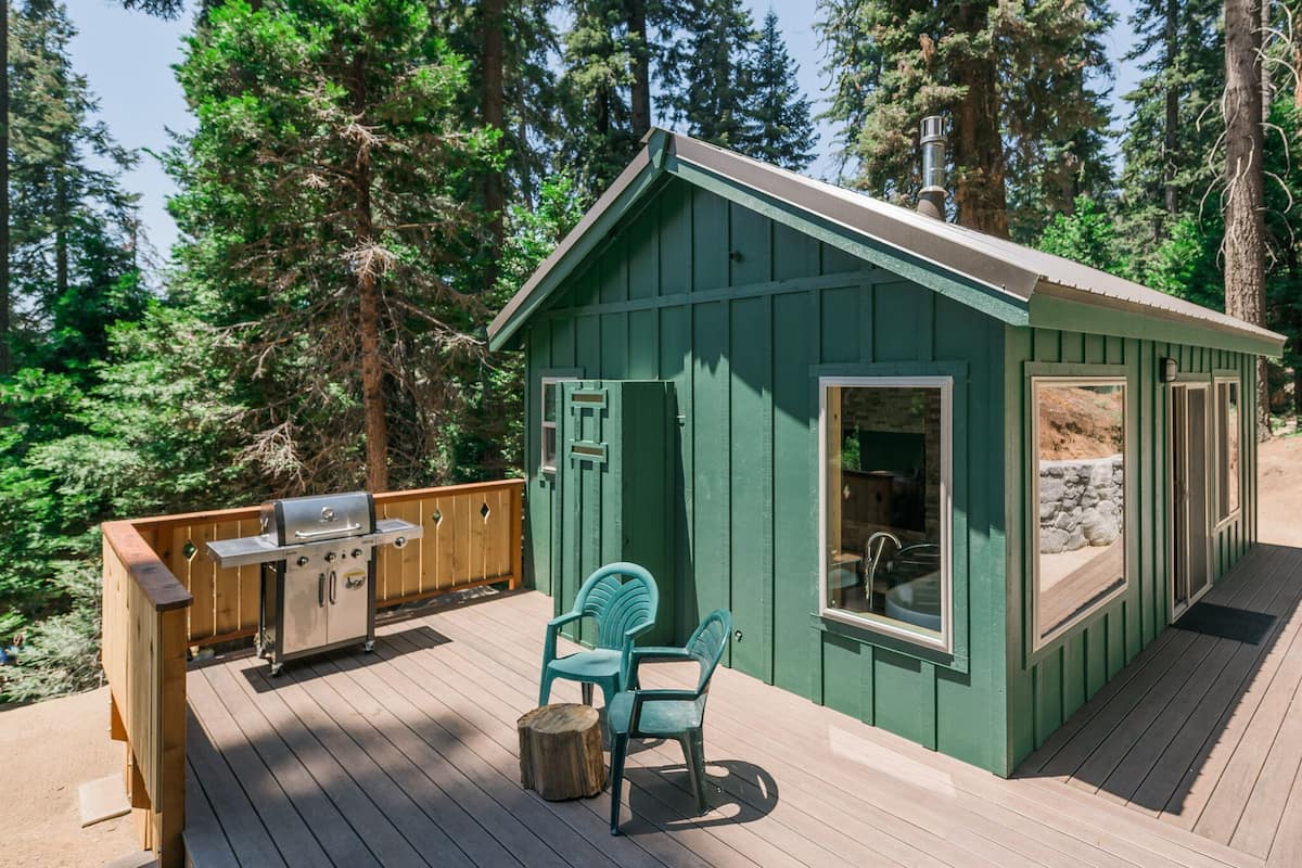 A green wooden cabin with a deck, a grill, and two chairs.