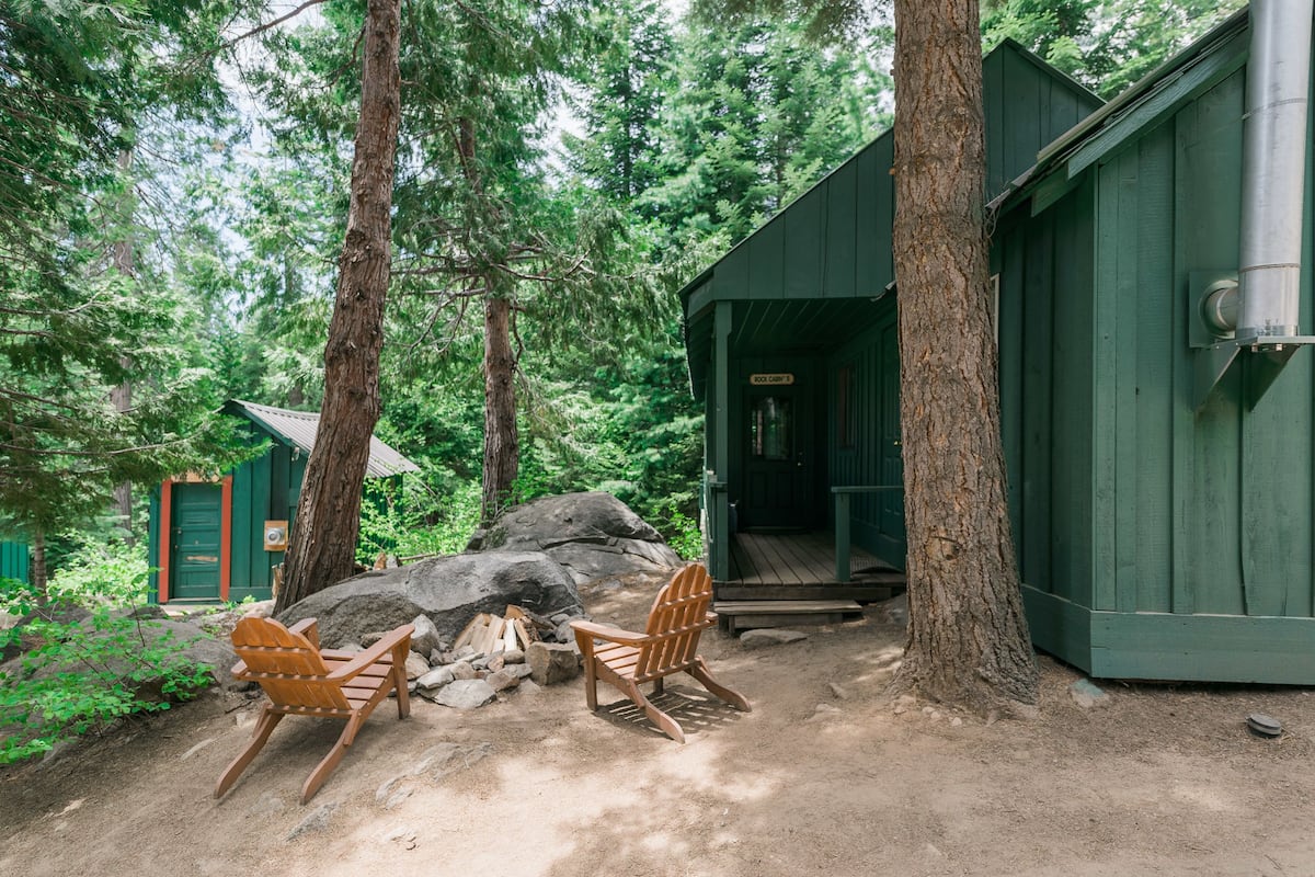 A cabin with a wooden deck, two rocking chairs, a fire pit, and a green building with a chimney.