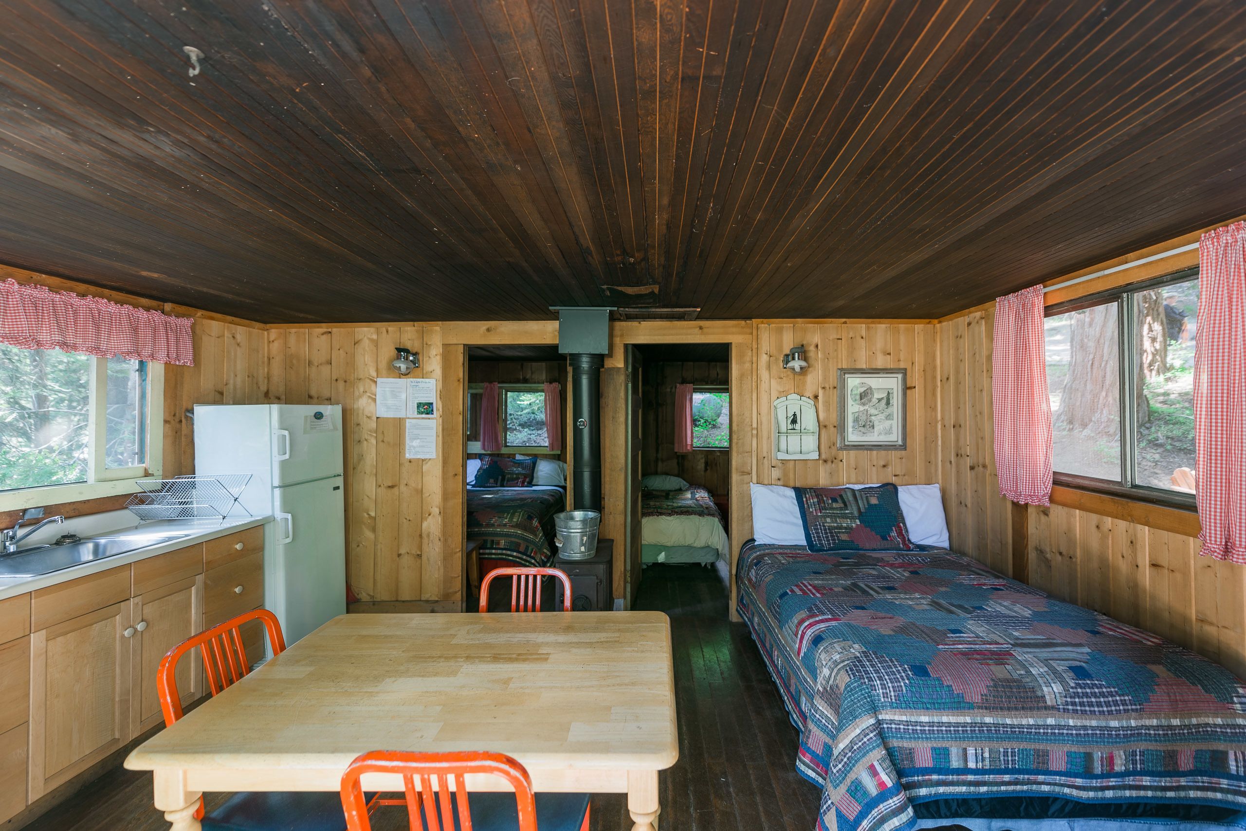 A cabin with a wooden interior, a dining table, two beds, a kitchen area, and a window with curtains.