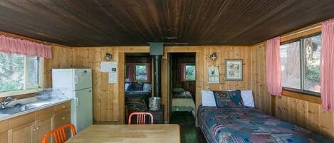 A cabin with a wooden interior, a dining table, two beds, a kitchen area, and a window with curtains.