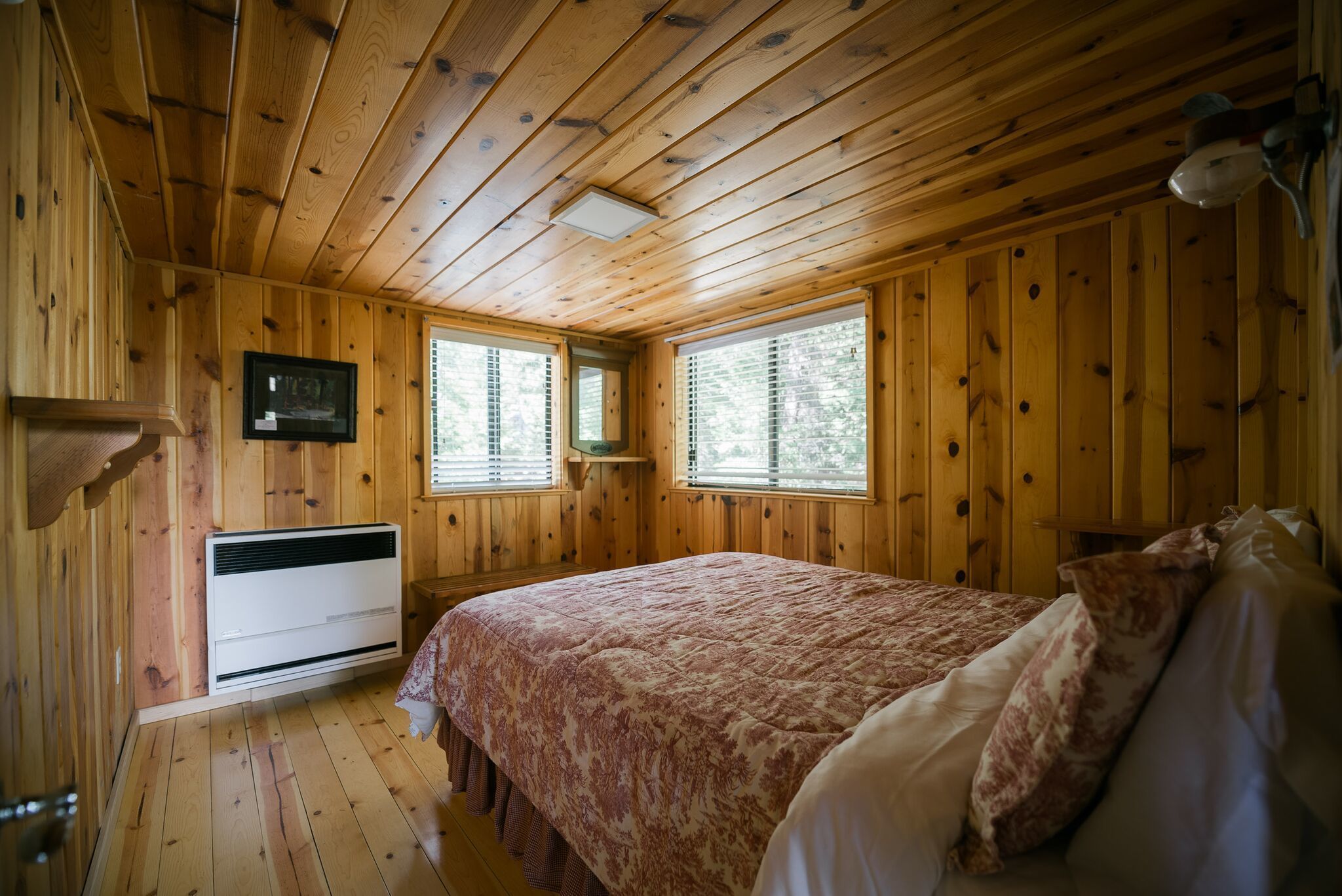 A wooden-paneled bedroom with a bed, a wall-mounted heater, and two windows with blinds.